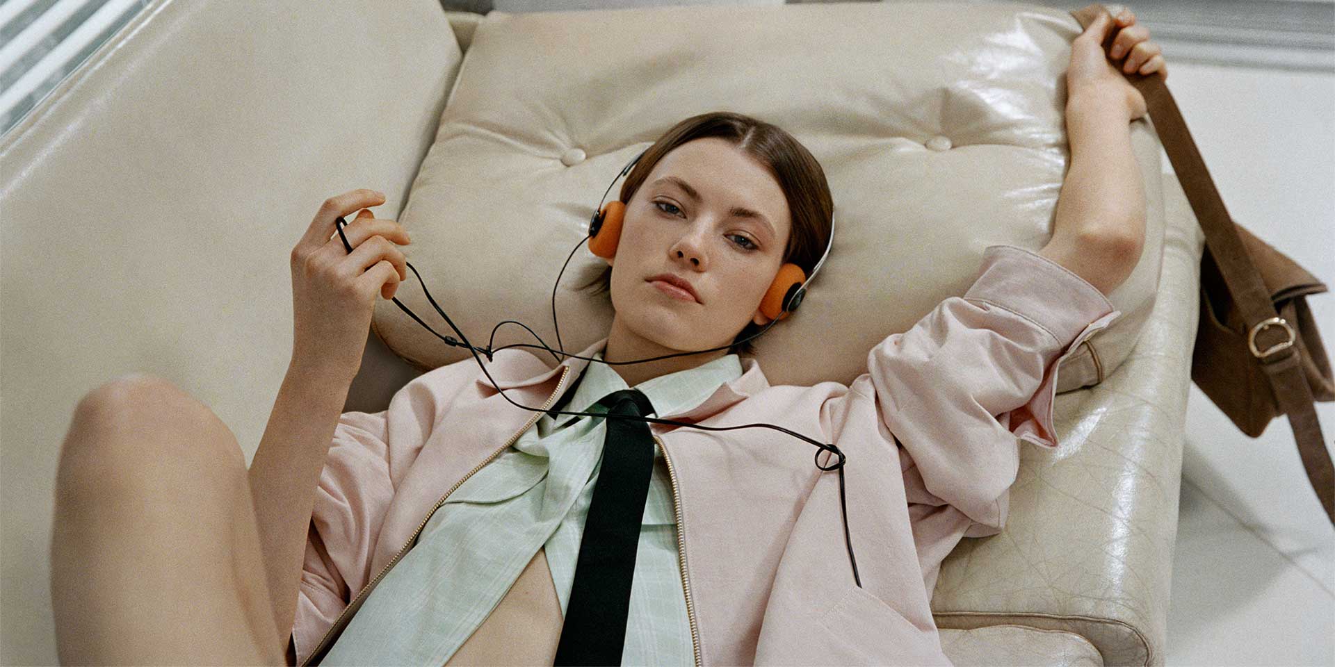 Dana Smith wearing headphones, a light pink coat, striped shirt, a black tie, and denim shorts, reclines on a beige sofa, advertising Sandro’s Spring–Summer 2026 campaign, photographed by Hart Lëshkina against modernist concrete architecture near Giverny.