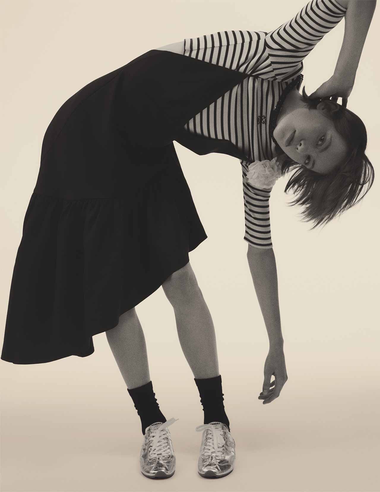 Dana Smith wearing a black Sandro dress with fluid silhouette and styled with silver Lightwave sneakers, SS26 editorial shot in a concrete modernist setting.